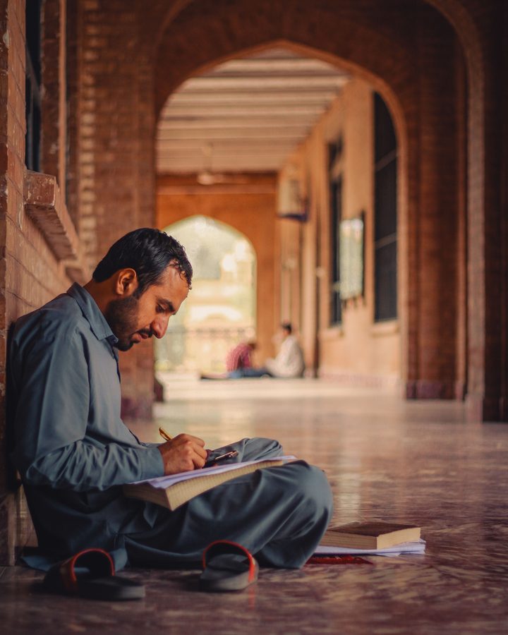 A person writing in a notebook along an arched stone corridor.